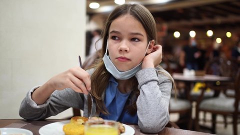 A teen girl with a protective medical mask on her chin eats a sausage in the hotel dining room. travelling and resting during a pandemic. 