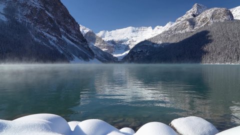Lake Louise in early winter sunny day morning. Mist floating on turquoise color water surface. Clear blue sky, snow capped mountains in background. Beautiful natural landscape in Banff National Park.