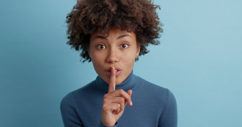 Shh be quiet please. Mysterious dark skinned Afro American woman presses index finger to lips makes silence gesture tells secret winks eye and wears casual poloneck poses against blue background