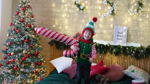 Little cheerful boy in an elf hat jumping on the bed with an inflatable cane in his hands