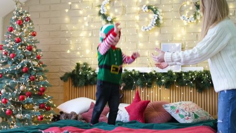 Little cheerful boy in an elf hat jumps on the bed holding his mother's hands