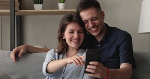 Married couple relax on sofa have fun using new cool free application on smartphone laughing enjoy lazy weekend with modern tech, watch videos on device or happy photo memories, carefree time concept
