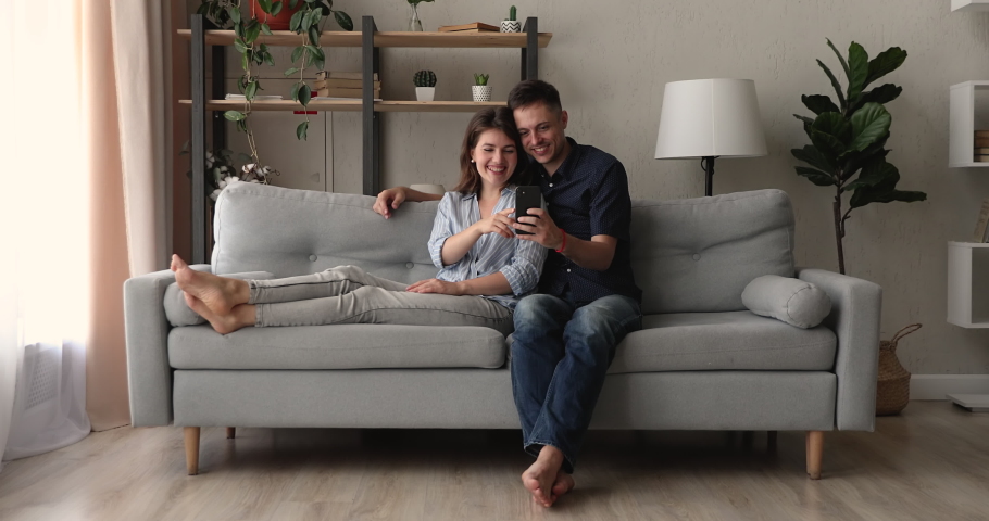 Millennial couple relaxing on couch taking selfie pictures using smartphone, having fun with new cool application, enjoy carefree weekend lazy day with wireless modern technology and internet concept | Shutterstock HD Video #1062745129