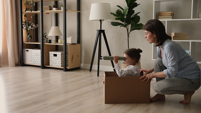 Mother pushes little daughter across the living room floor in a cardboard box. Royalty-Free Stock Footage #1062745144