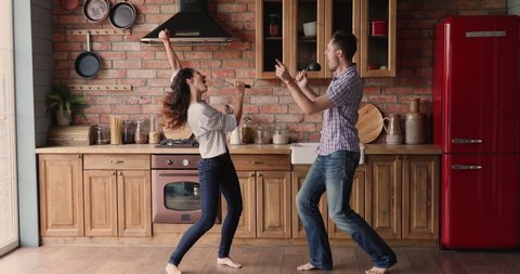 Cheerful girlfriend and boyfriend listen music dancing barefoot in kitchen sing song use kitchen utensil like microphone in microphones feels happy. Funny weekend activity, home karaoke, hobby concept