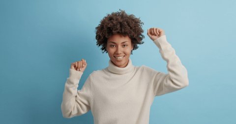 Joyful curly haired young woman makes champion dance celebrates good news raises arms enjoys favorite music dressed in white jumper isolated over blue studio background feels empowered and free