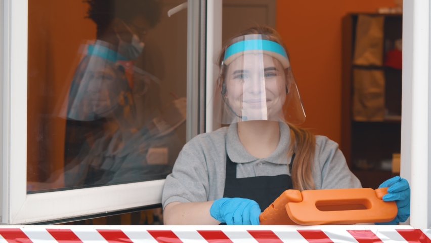Fast food cashier with tablet and headset in drive thru service wearing face shield and glasses smiling at camera. Pretty cafe staff in delivery window looking at client
