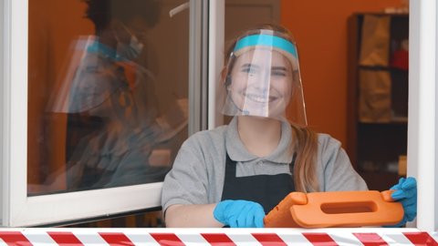 Fast food cashier with tablet and headset in drive thru service wearing face shield and glasses smiling at camera. Pretty cafe staff in delivery window looking at client