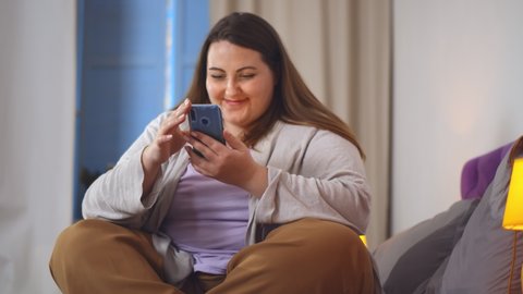 Portrait of obese smiling woman sitting cross-legged on bed and chatting on smartphone. Young overweight female surfing internet on mobile phone relaxing in bedroom at home