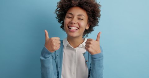 Lovely cheerful ethnic woman makes like gesture raises thumb up and appreciates something nice smiles broadly being in good mood poses against blue studio wall. I really recommend this to you