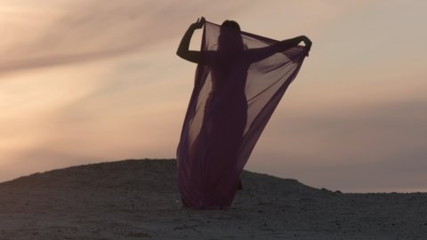 A beautiful woman in an oriental costume walks on the sand at sunset. Raises a transparent veil overhead that flutters in the wind. Shooting from behind. Silhouette.