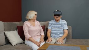 Enthusiastic grandfather and grandmother with VR headset helmet play games, watch virtual reality 3D 360 video. Family of senior man and woman at home. Future technology. VR goggles, glasses