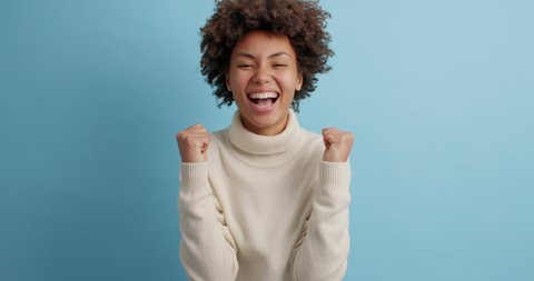 Overjoyed curly haired woman wins competition achieves goal and makes triumph gesture says yes feels like winner shows fist bump happy dream come true wears white jumper poses against blue wall