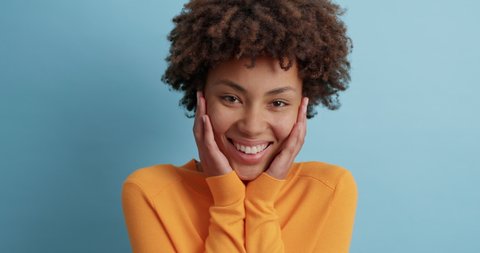 Sincere positive Afro American woman laughs out loudly doesnt hide emotions keeps hands on cheeks and smiles broadly feels happiness dressed in casual jumper isolated over blue studio background