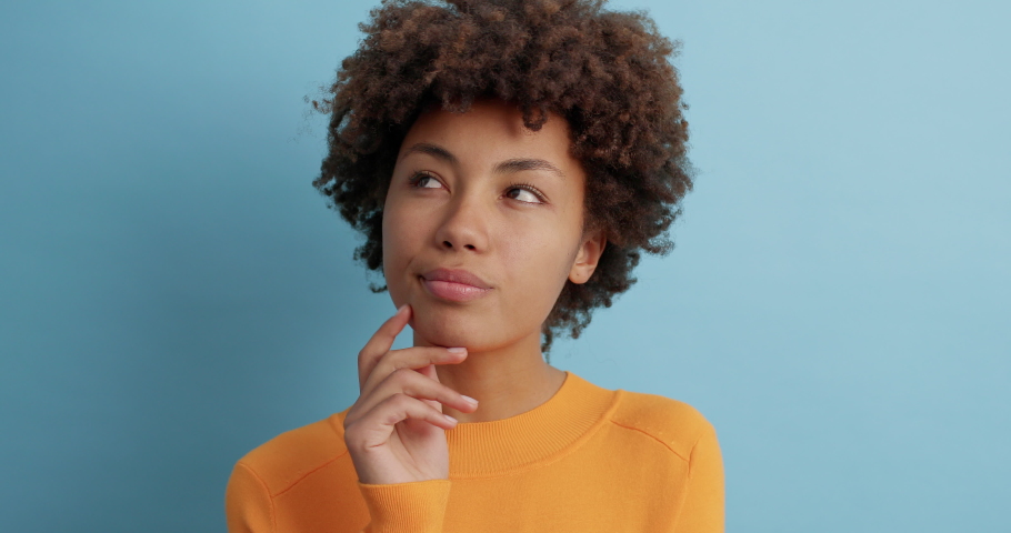 Thoughtful young Afro American woman looks somewhere pensively thinks deeply about something tries to make decision in mind wears casual orange jumper poses against blue background. Let me think | Shutterstock HD Video #1062750388