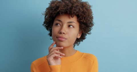 Thoughtful young Afro American woman looks somewhere pensively thinks deeply about something tries to make decision in mind wears casual orange jumper poses against blue background. Let me think