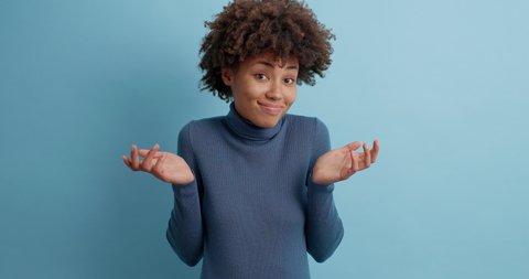 Pretty curly woman shrugs shoulders unaware raises hand clueless has no idea cannot answer wears casual turtleneck poses against blue background. Ethnic beautiful girl not knowing information