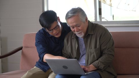 happy asian young son and senior old father sitting on sofa using laptop computer together at home