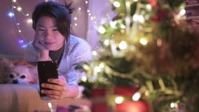 asian female woman happiness cheerful hand hold smartphone video call to her friend laydown on sofa with cute lovely puppy little dog in christmas holiday vacation festive background concept