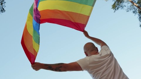 Queer man holding rainbow gay flag while parade on background of blue sky. Happy guy wearing heart sunglasses demonstrate his rights. LGBTQI, Pride Event, LGBT Pride Month, Gay Pride Symbol Video de stock