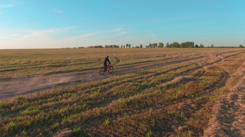 Man in green helmet rides orange enduro electro full suspension mtb mountain electric bicycle e-bike along rural dirty road dust from wheels past field wood on sunny autumn sunset evening aerial view.