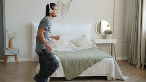 Slow motion of happy guy in pajamas dancing in bedroom wearing wireless headphones having fun alone. Modern lifestyle and creative youth concept.