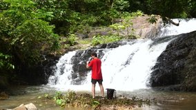 Back View of Traveler Man in Red T-shirt Enjoying Beautiful Breathtaking Waterfall Using Smartphone and Shooting Photo or Video in Thailand Rainforest. Great Wild Jungle Nature