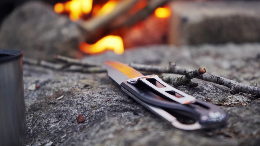 A small sharp camping knife lies on a stone in front of a burning bonfire. Slow motion footage.