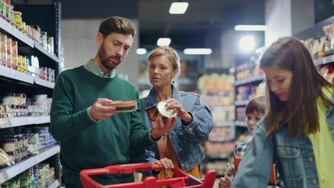 Affectionate young beautiful family of four people choosing product items on shelves shopping together inside modern supermarket.