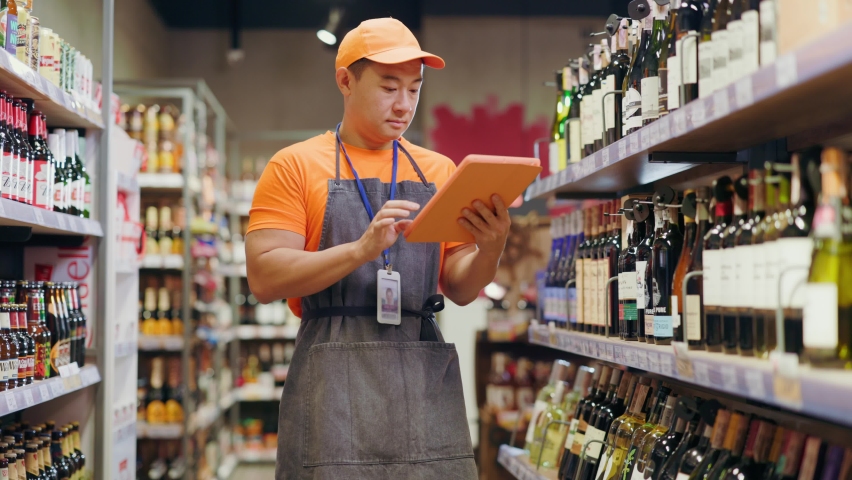 Young adult chinese salesman employee workman using tablet computer management controlling quality and number of alcohol bottles in grocery store. Royalty-Free Stock Footage #1062772492