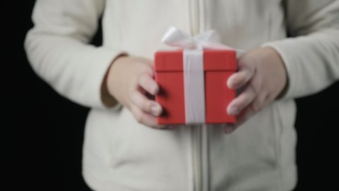 closeup on little girl's hands giving red box with white ribbon. winter holidays, christmas, new year, surprise. small child gives present in box on black background. Person holds gift for birthday