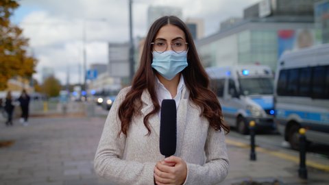 Caucasian Confident Young Woman Journalist In Protective Mask With Microphone Presenting Breaking News TV About Coronavirus Pandemic On Street Outdoors Cars On Signals Police In Background
