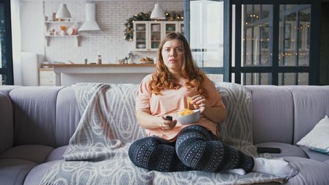 Couch potato. Young overweight lady sitting on sofa at home, watching tv and eating junk chips