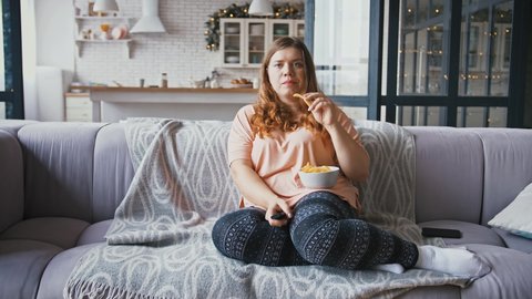 Young overweight woman sitting on couch at home, watching tv and eating potato chips