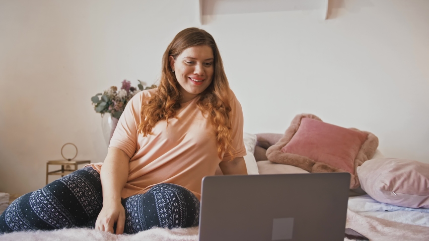 Online communication. Young plus size lady talking with friends via video call on laptop, sitting on bed at home Royalty-Free Stock Footage #1062774403