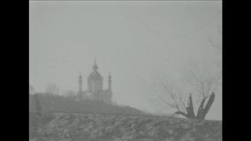 
a man in a jacket with a hood gulet with a dog on a leash. against the background of an ancient church, with domes. 16 mm shot on a rare film camera on black and white film.