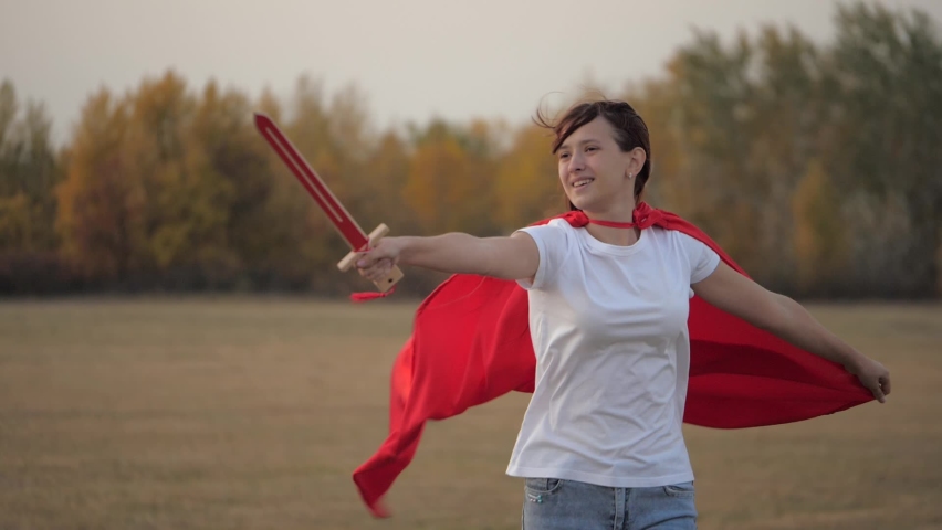 Child plays spartnats. a free girl in a red cloak with a sword in her hand plays a medieval knight in the sun. young girl plays superheroes. girl fights with a toy sword. happy childhood concept.