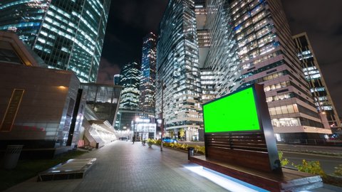 Modern billboard with a green screen on a busy street of the big city in the night. Road traffic and gigantic skyscrapers glittering with neon lights and pedestrians passing by in motion timelapse.