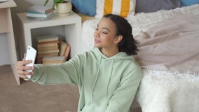 Happy generation z african american teen girl influencer blogger using mobile cell phone, taking selfie for social media apps, chatting with friends online on smartphone sitting in bedroom at home.