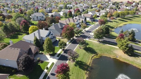 Overhead aerial view of colorful autumn trees, residential houses and yards with drainage pond along suburban street in Chicago area. Midwest USA. 4K