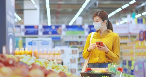 asian young businesswoman wearing face mask is shopping for fresh organic fruits and check shopping list with smart phone at supermarket during virus epidemic outbreak