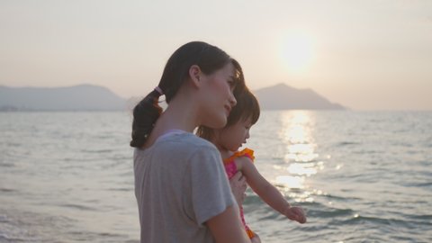 Slow motion footage of happy Asian mother standing on the beach holding her baby girl in arm. She pointing in the sky talking to her kid. Mom kiss daughter in beautiful twilight sky in the evening.