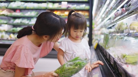 Young Asian beautiful mother holding grocery basket with her child walking in supermarket. She is choosing green salad vegetable picking up from shelf and give it to her little kid. Healthy shopping.
