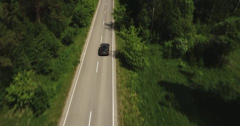 One black SUV car driving alone, travel on empty straight asphalt road freeway through dense forest corridor at summer sunny day. Aerial drone wide view.