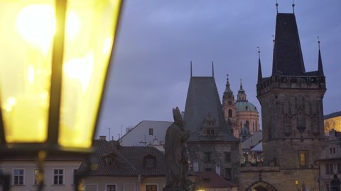 Night illuminated Charles Bridge with gas streetlight lantern against towers and facades of ancient architectural buildings with large spires under cloudy sky in evening