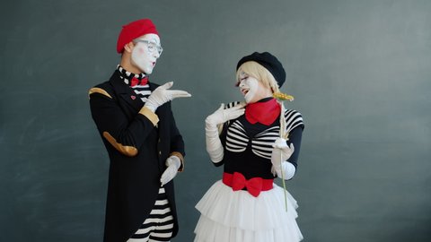 Man mime is giving flower to beautiful girl getting air kiss and thumbs-up hand gesture standing on grey background. People and relationship concept.