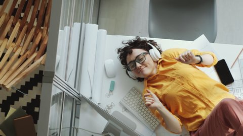 Top down shot of young businesswoman in headphones lying on floor plans on office desk, smiling and dancing while listening to music with eyes closed