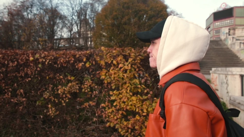 Guy in cap and hood on his head moves through city park. Walking around city. Using backpack to carry things. Camera movement.