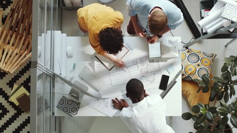 Top down shot of group of multiethnic male and female architects unrolling paper on office table, discussing construction plan and putting marks while working in team on project