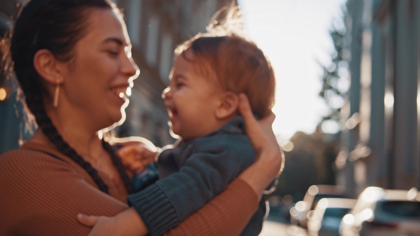 Joyful and cute young mom hold little child in her arms, hug and having fun together. Happy childhood and motherhood concept. Laughing, smiling, emotions, enjoy weekend outdoors, sunny city street Royalty-Free Stock Footage #1062802138